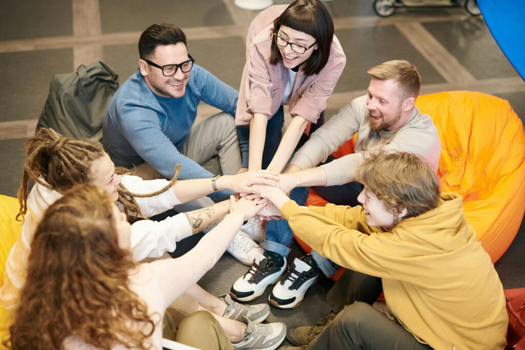 A diverse group of adults sit indoors, joyfully stacking hands together, symbolizing teamwork and camaraderie.